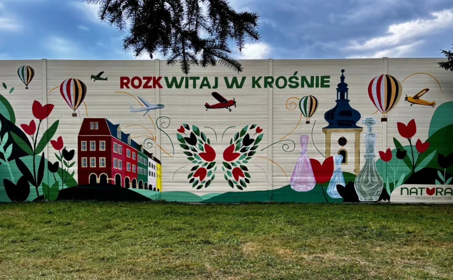 Mural na jasnym, podzielonym na segmenty murze z napisem „ROZKWITAJ W KROŚNIE”. Malowidło przedstawia charakterystyczne motywy miasta, w tym kolorowe kamieniczki, wieżę kościelną, szklane naczynia, balony na ogrzane powietrze oraz samoloty, z centralnie umieszczonym motywem motyla złożonego z liści i kwiatów.